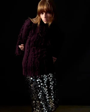 Model wearing a dark purple fringed sweater and metallic sequin skirt against a solid black studio background.