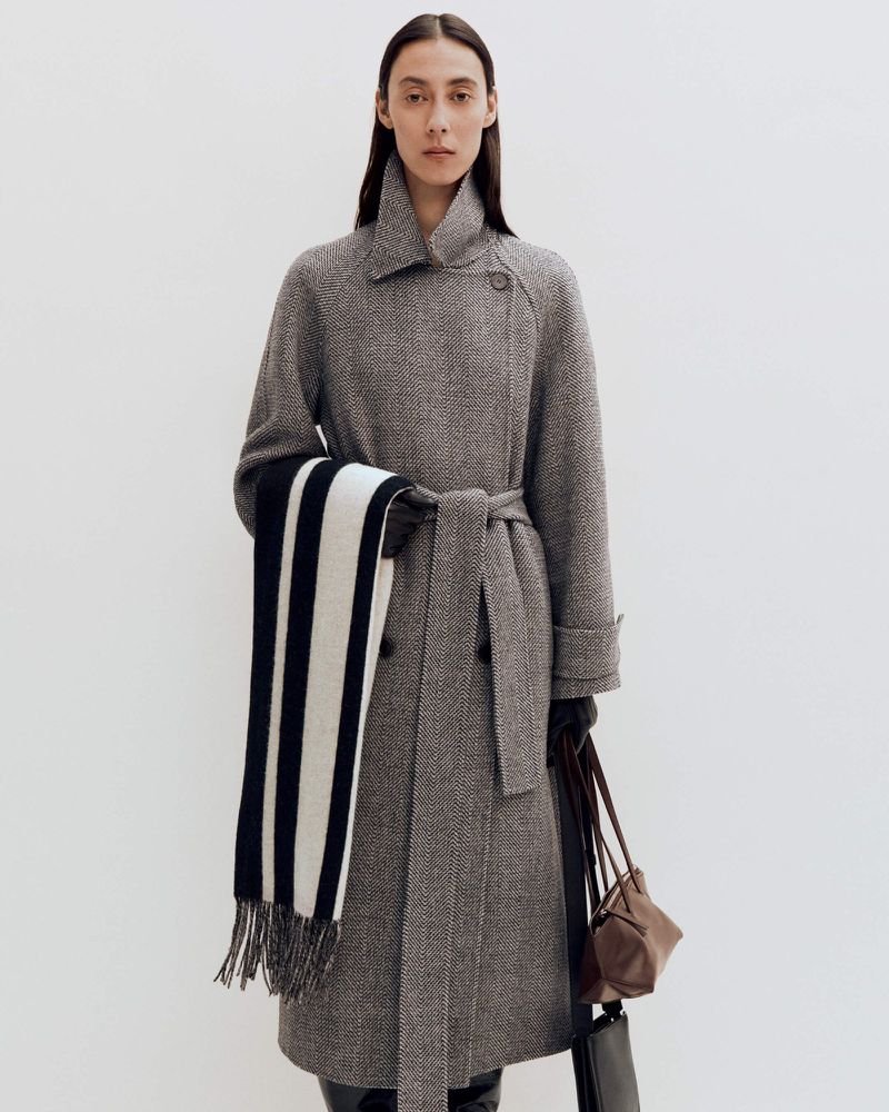 A woman in a herringbone wool coat with a belted waist and scarf, holding a brown leather purse, posing against a white background.
