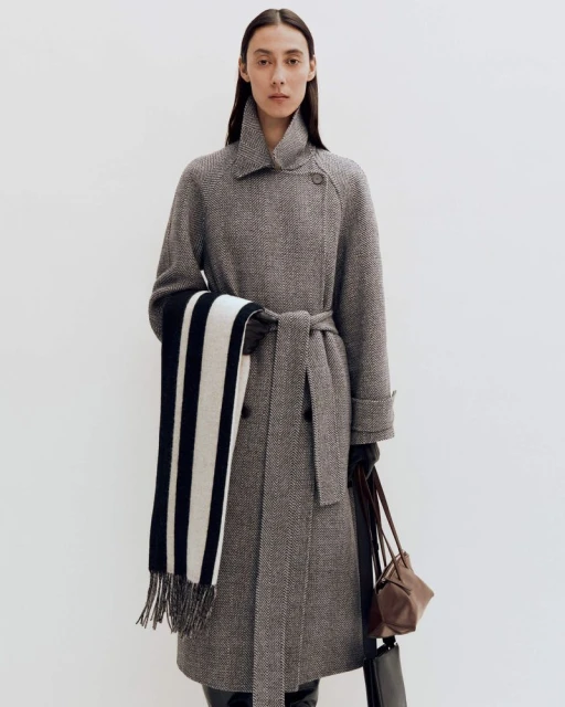 A woman in a herringbone wool coat with a belted waist and scarf, holding a brown leather purse, posing against a white background.