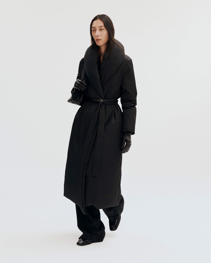 Woman in a long black puffer coat with a belt and a black purse in a bright studio setting, showcasing a modern, fashionable outerwear look.