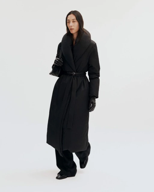 Woman in a long black puffer coat with a belt and a black purse in a bright studio setting, showcasing a modern, fashionable outerwear look.