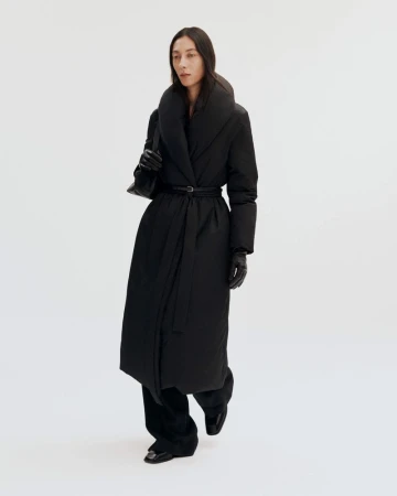 Woman in a long black puffer coat with a belt and a black purse in a bright studio setting, showcasing a modern, fashionable outerwear look.
