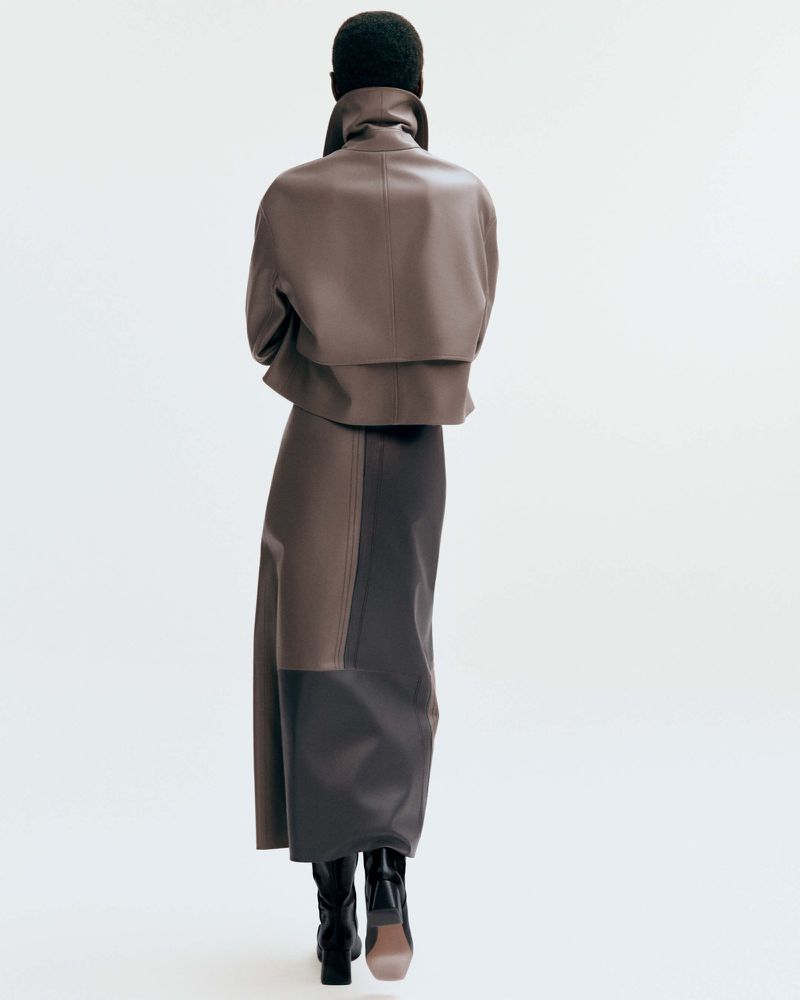 A slender adult fashion model is viewed from behind in a brown leather jacket and a color-blocked skirt against a white backdrop.