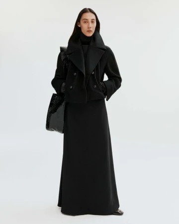 Fashion shot of a woman in a black wool coat and long skirt holding a shiny black tote bag in a bright white studio setting.