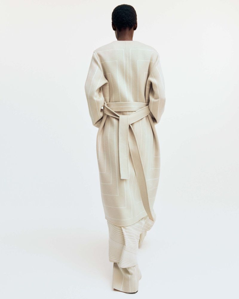 A slender, dark-haired person stands facing away, modeling a patterned cream-colored coat with a matching sash and loose-fitting trousers.