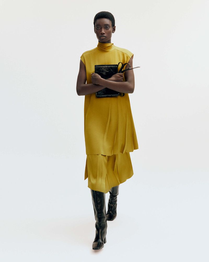 Fashion model in a mustard dress and black boots holds a patent leather bag against a white background in a studio setting.