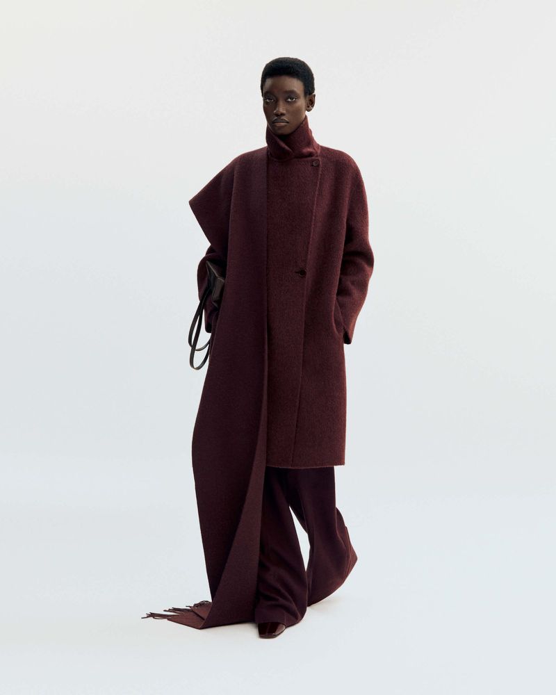 A dark-skinned woman poses in a dark red, oversized wool coat and matching pants against a white background in a studio setting.