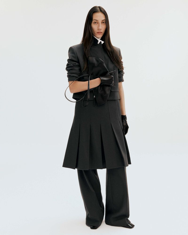 A long-haired woman poses in a gray skirt, jacket, and gloves on a white background, holding a purse with a serious expression.