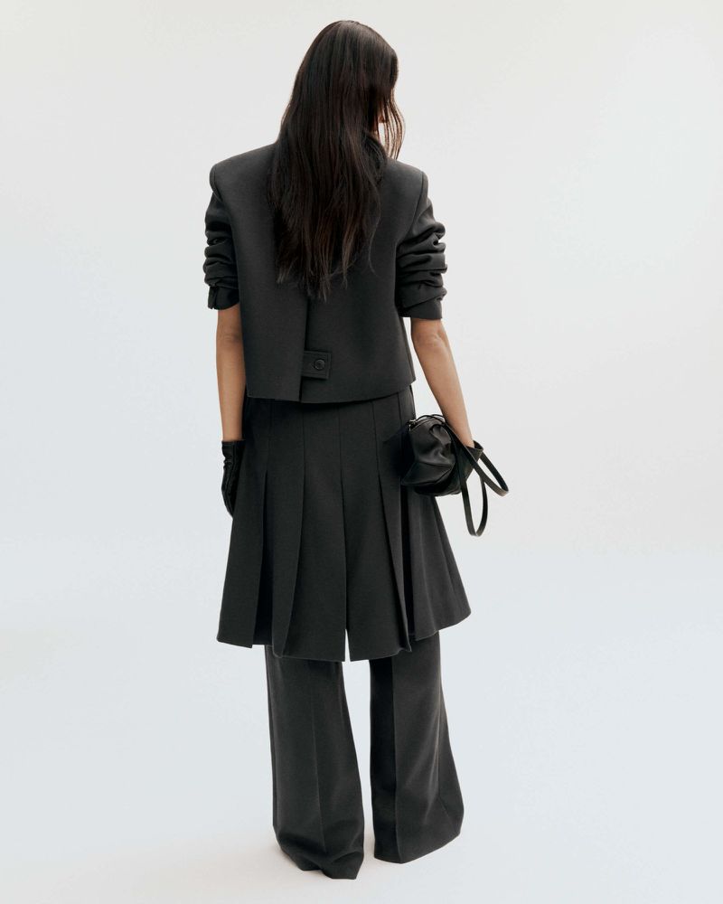 Fashion shot featuring a woman in a dark grey crop jacket, pleated skirt, wide leg pants, gloves, and a small purse against a white backdrop.