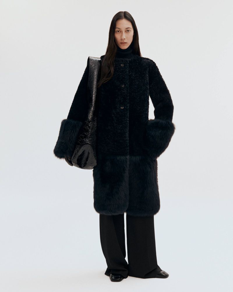 Model wearing a dark textured coat and carrying a large black shoulder bag against a plain white studio background.