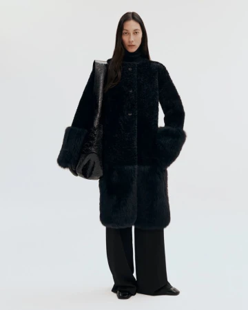 Model wearing a dark textured coat and carrying a large black shoulder bag against a plain white studio background.