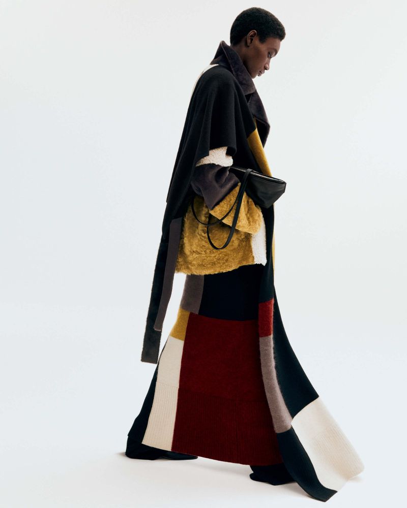 Editorial fashion shot of an African-descent adult female in a long, colour-blocked coat with fur bag, posing against a white backdrop.