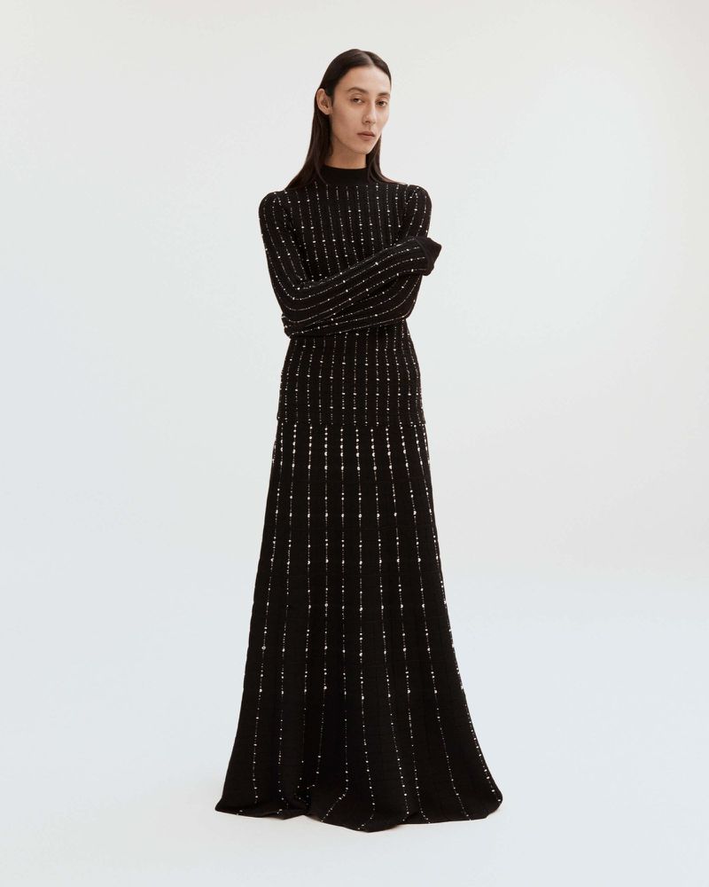 Elegant, full-length shot of a slender female model posing in a black, floor-length knit dress with subtle metallic detailing against white background.
