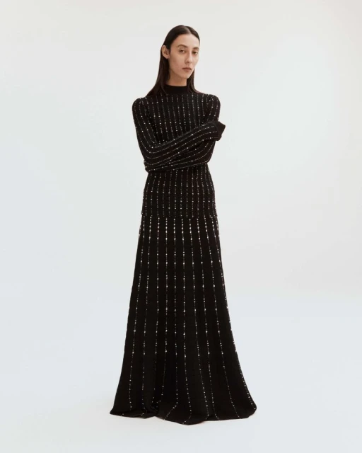 Elegant, full-length shot of a slender female model posing in a black, floor-length knit dress with subtle metallic detailing against white background.