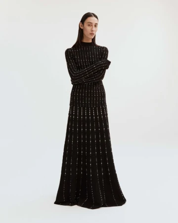 Elegant, full-length shot of a slender female model posing in a black, floor-length knit dress with subtle metallic detailing against white background.
