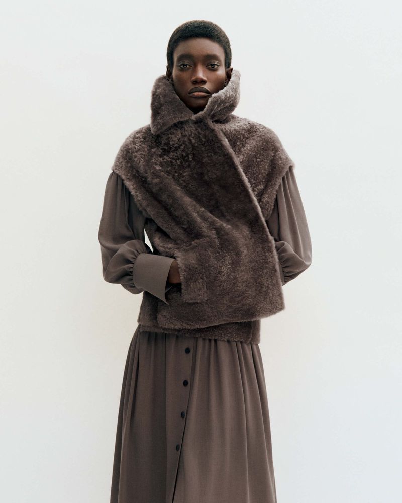 Dark-skinned woman wears a long-sleeved gray dress and a faux fur shawl against a stark white background in a full-body studio portrait.