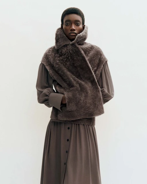 Dark-skinned woman wears a long-sleeved gray dress and a faux fur shawl against a stark white background in a full-body studio portrait.