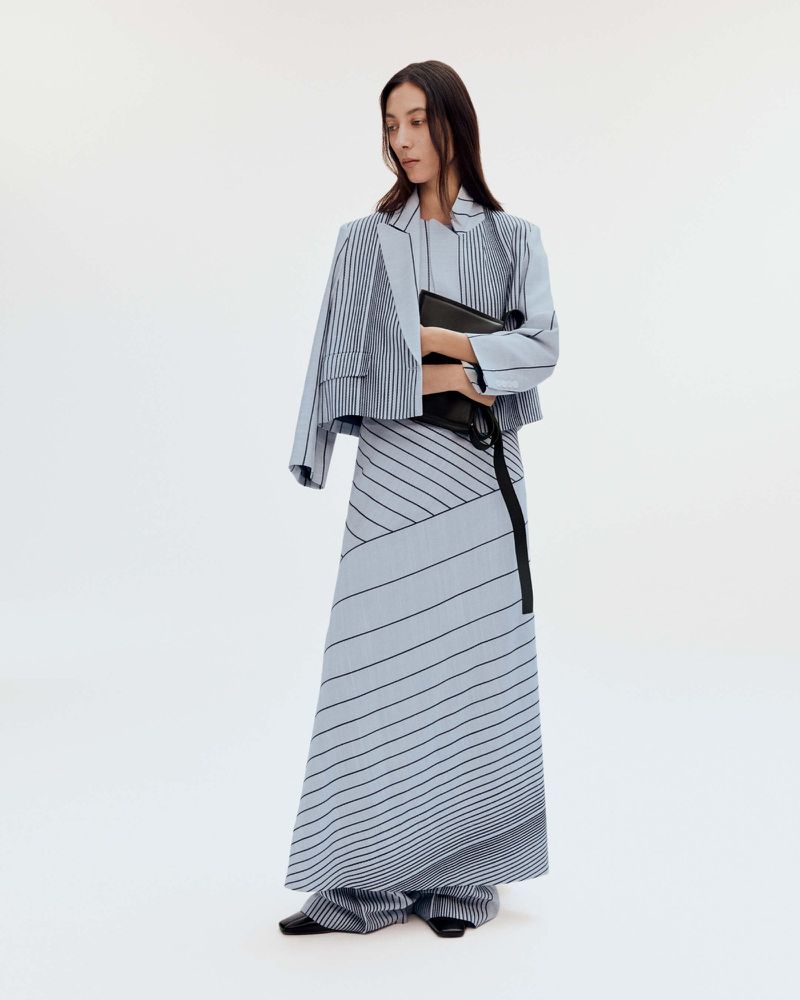 Elegant adult woman in light blue striped blazer and long skirt holding a black clutch bag in a bright, clean studio setting.