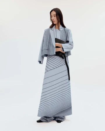Elegant adult woman in light blue striped blazer and long skirt holding a black clutch bag in a bright, clean studio setting.