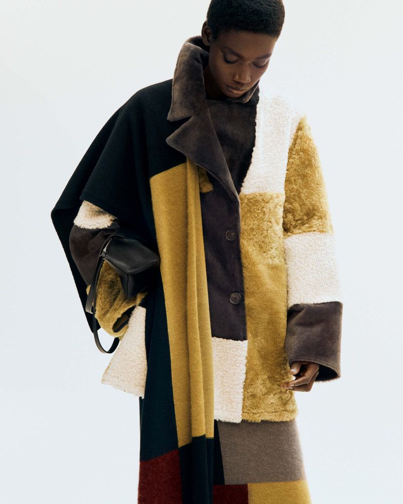 An African American woman models a patchwork coat and skirt in a fashion editorial shot against a stark white backdrop in a studio.