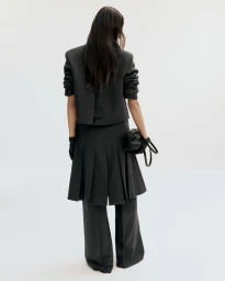 Model from behind wearing a black blazer, pleated skirt over trousers, and holding a black handbag against a white background