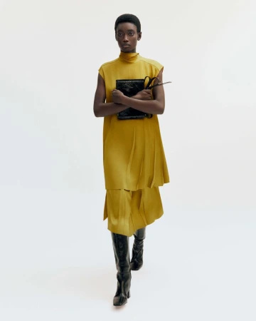 Model in mustard yellow sleeveless tunic holding a black handbag against a white background