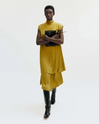 Model in mustard yellow sleeveless tunic holding a black handbag against a white background
