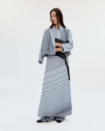 Model standing against a white seamless background wearing a blue striped suit jacket and matching long skirt, holding a black clutch.