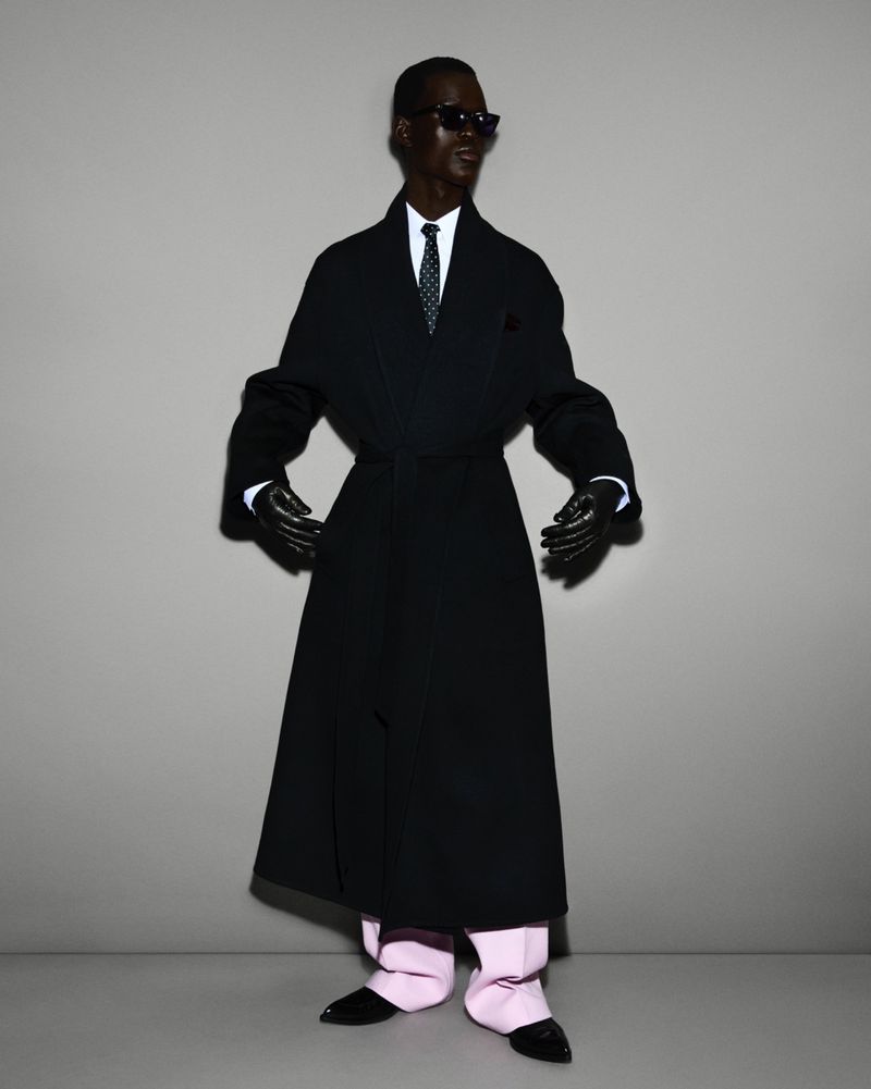 Fashion shot of dark-skinned adult male in long dark coat, white shirt and tie, light pink trousers, black gloves and shoes on grey backdrop.