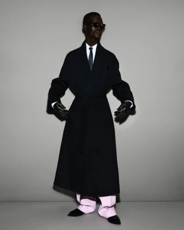 Fashion shot of dark-skinned adult male in long dark coat, white shirt and tie, light pink trousers, black gloves and shoes on grey backdrop.