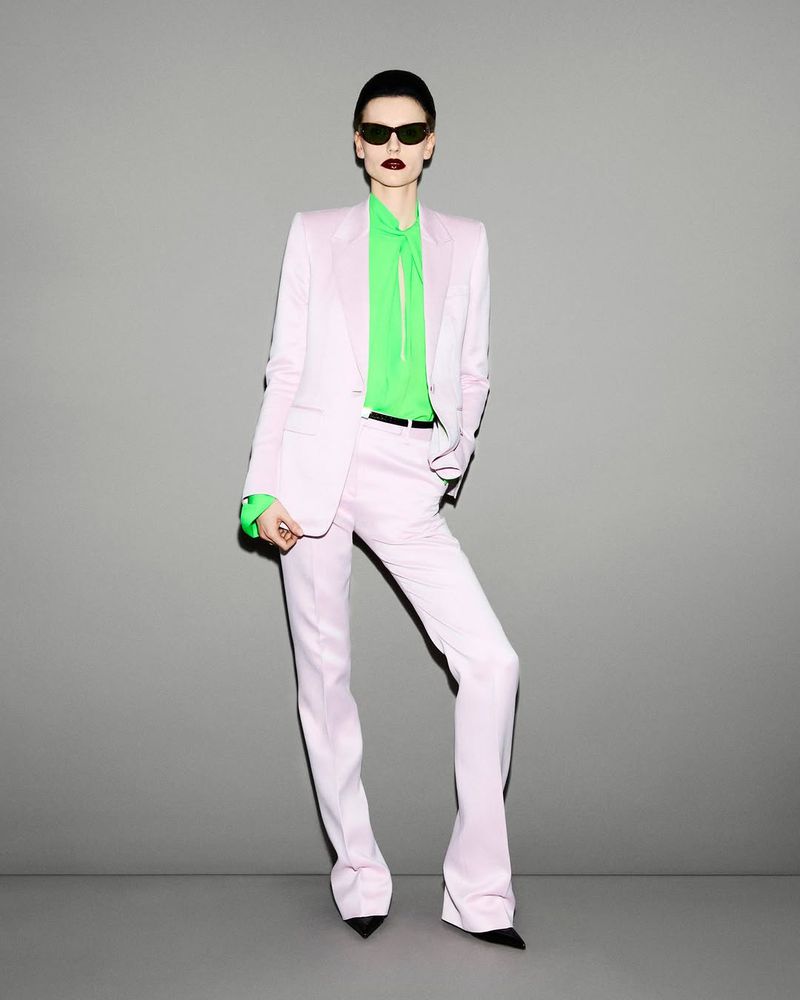 Fashion-forward woman in a pale pink suit and green top, posing against a neutral grey backdrop, highlighted by sunglasses and sleek styling.