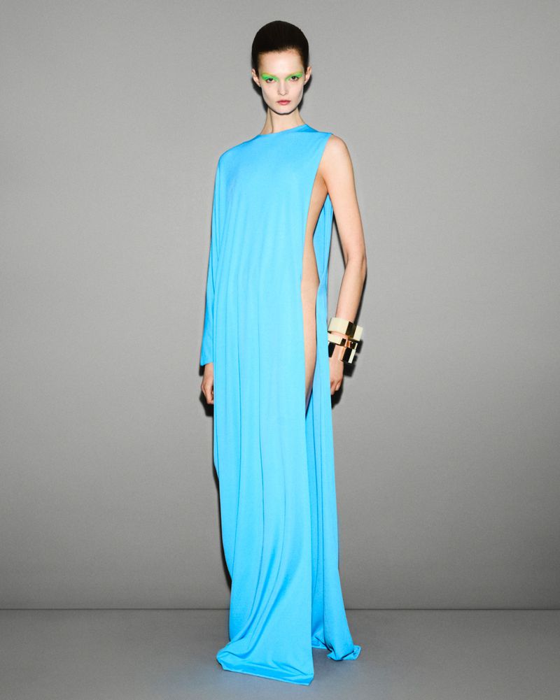 Elegant adult woman in a floor-length turquoise gown with one sleeve, posing against a gray studio backdrop for a fashion editorial.
