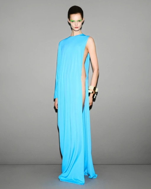 Elegant adult woman in a floor-length turquoise gown with one sleeve, posing against a gray studio backdrop for a fashion editorial.