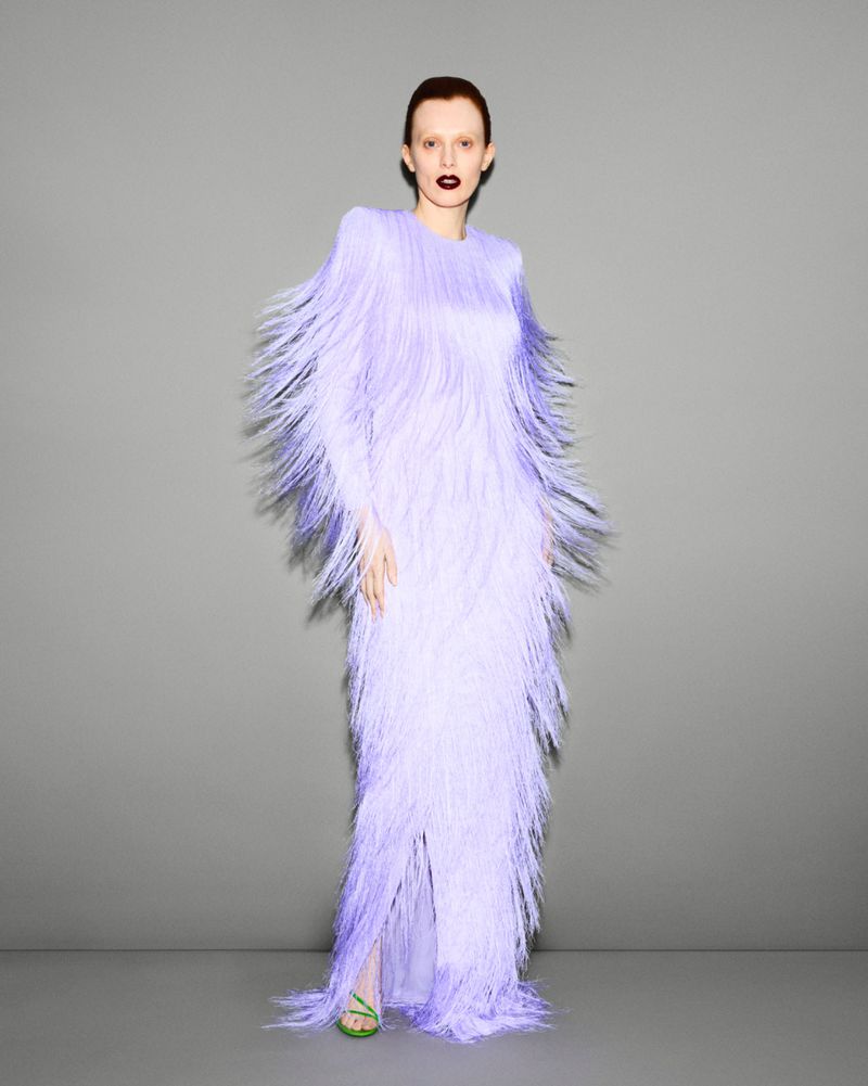 Elegant woman in lavender gown with fringe detail, posing in front of a neutral gray backdrop for a fashion editorial portrait.