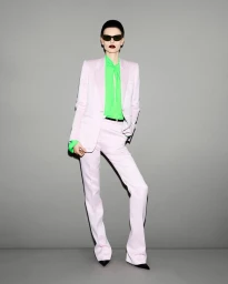 Model wearing a light pink suit, bright green blouse, and dark sunglasses against a plain grey background