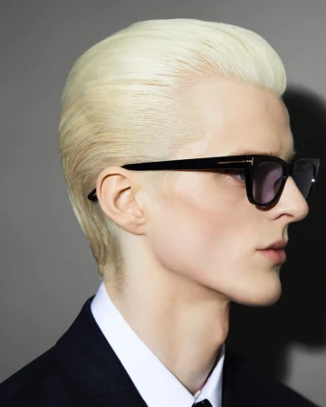 Close-up profile of a blonde model wearing black sunglasses and a dark suit jacket against a grey background.