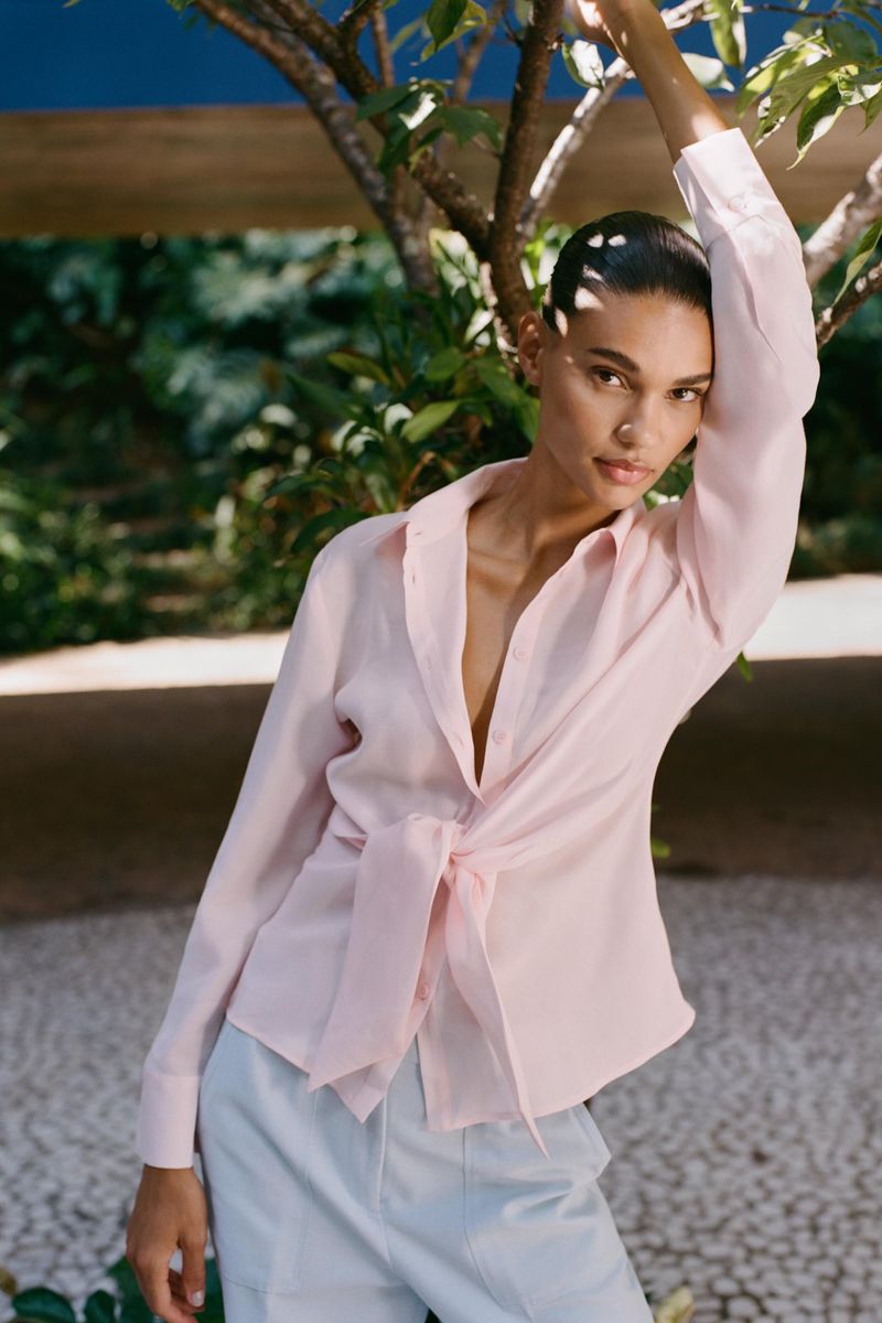 Adult female fashion model poses outdoors wearing pink button-down shirt and light blue pants, greenery and foliage in the background.