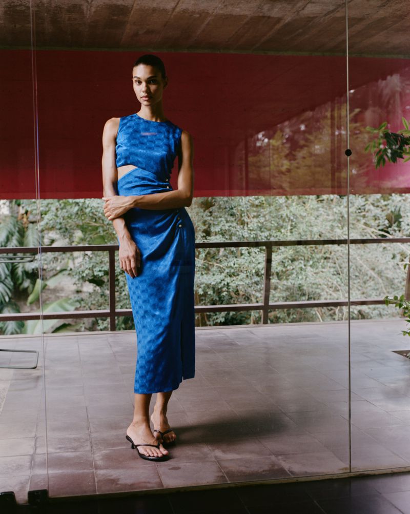 A young adult woman poses in a blue patterned sleeveless dress with a side cut-out in a sunlit indoor-outdoor architectural space.