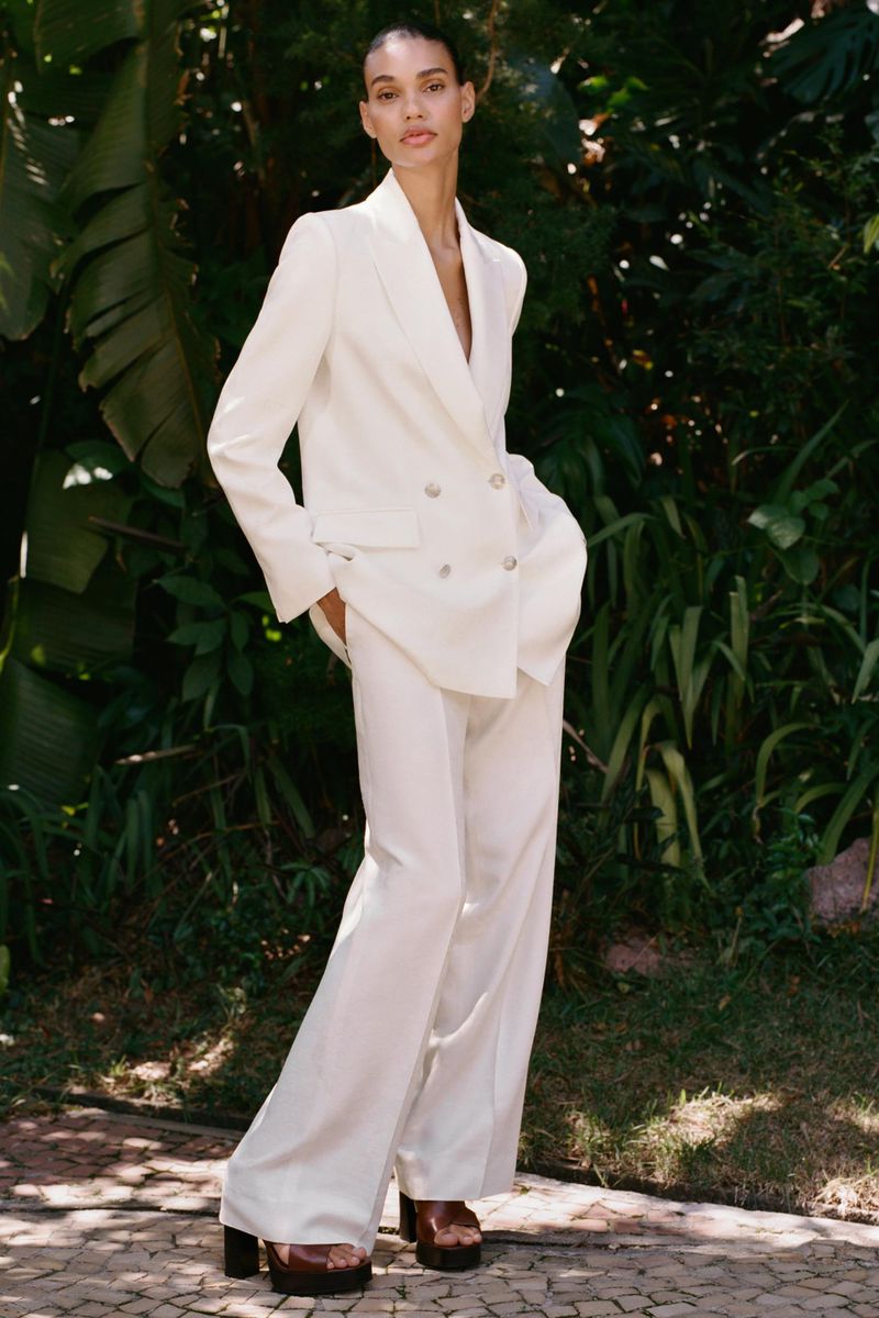 A woman in a white suit stands in a garden with lush greenery, wearing a double-breasted blazer and flared trousers, posing outdoors.