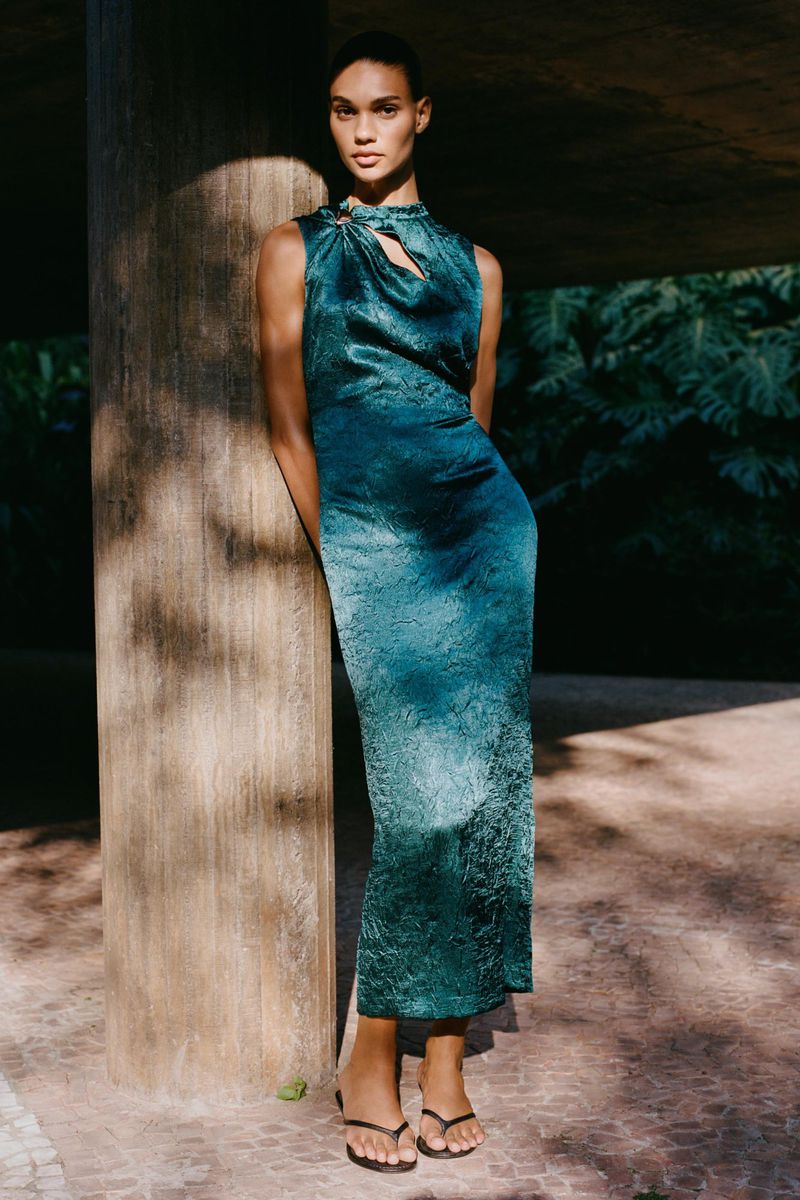 Elegant adult woman poses in a teal sleeveless dress against a weathered pillar, with lush greenery in the soft-lit background.
