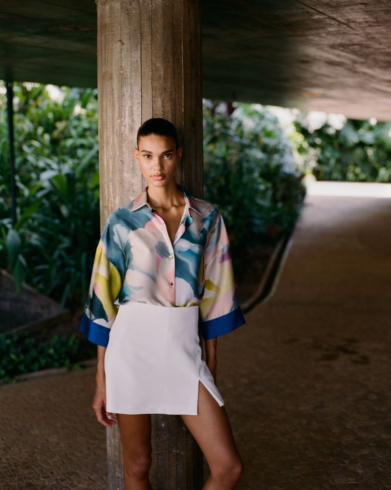 An adult woman in a patterned blouse and white skirt stands by a concrete pillar in a semi-outdoor space with green foliage.