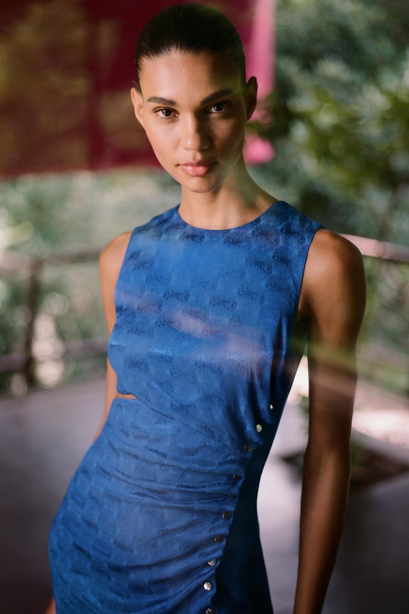 Elegant adult female model in blue sleeveless dress with side cutout poses in outdoor setting with greenery visible in background.