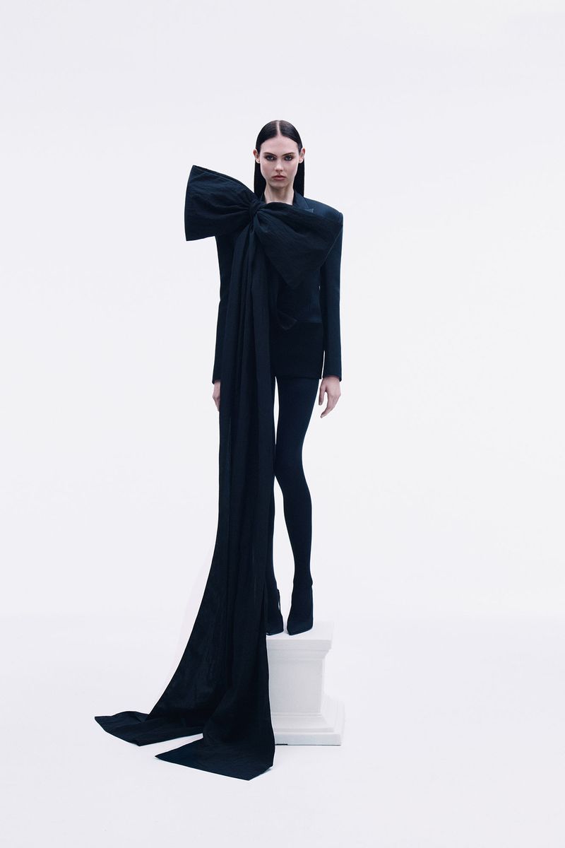 A woman in a black suit with a large bow detail stands on a pedestal in a stark white photo studio, fashion-forward and minimalist.