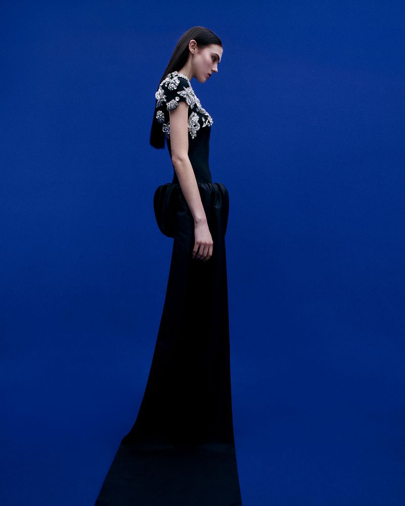 Model in a black embroidered gown standing against a solid blue background for David Koma Autumn-Winter 2025.