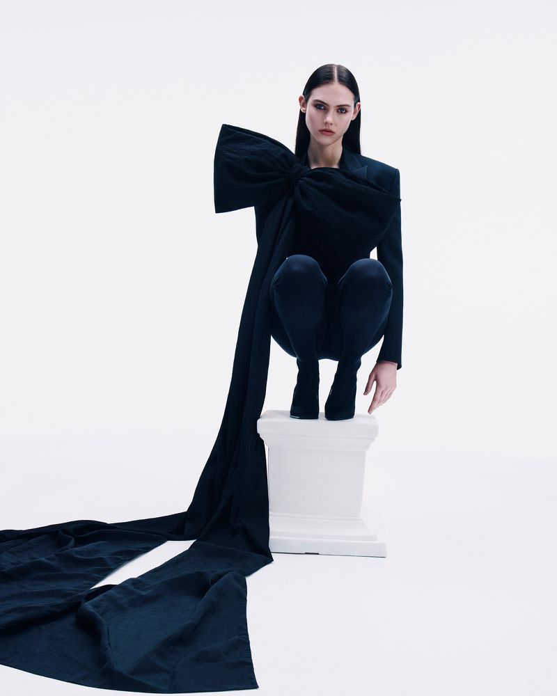 Woman in a dark fashion garment with an oversized bow poses on a pedestal, standing out against a white background in a studio setting.