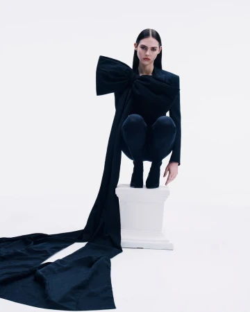 Woman in a dark fashion garment with an oversized bow poses on a pedestal, standing out against a white background in a studio setting.