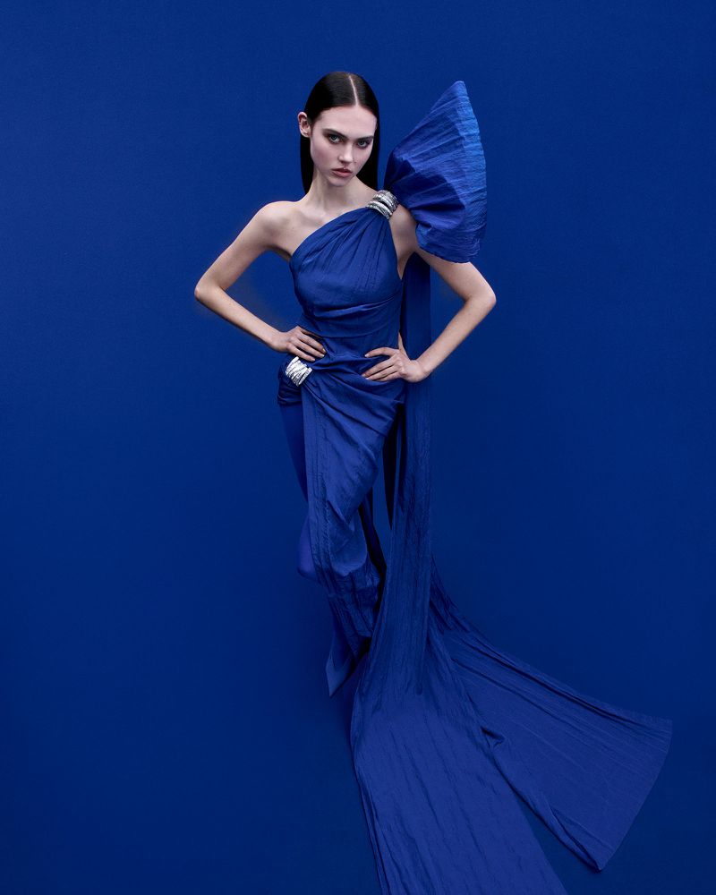Fashion shot of a woman in a draped, one-shoulder blue gown with a large bow detail, hands on hips, against a solid blue background.