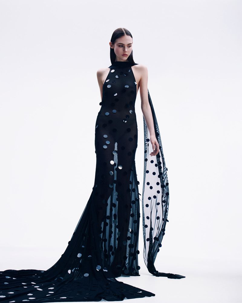 A slender model showcases a full-length black halter gown adorned with large polka dots, complete with a draped polka dot shawl, against a white backdrop.
