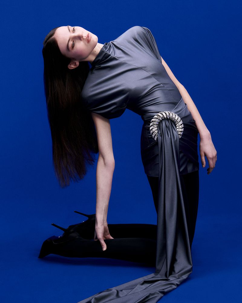 Fashion photo of a woman in a grey dress kneeling against a blue backdrop. The dress features a decorative embellishment at the waist.