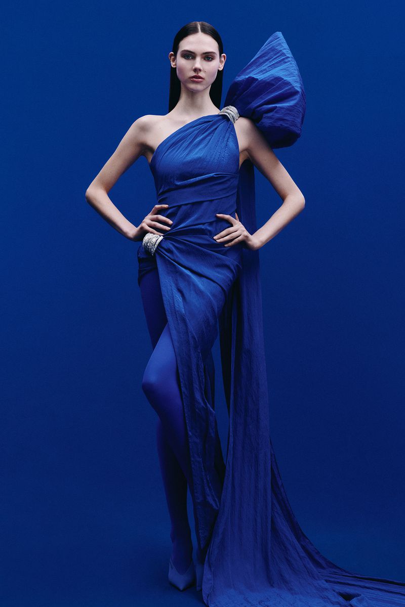 A slender woman poses in a one-shoulder blue gown with a large bow, set against a matching blue backdrop in a fashion photography style.